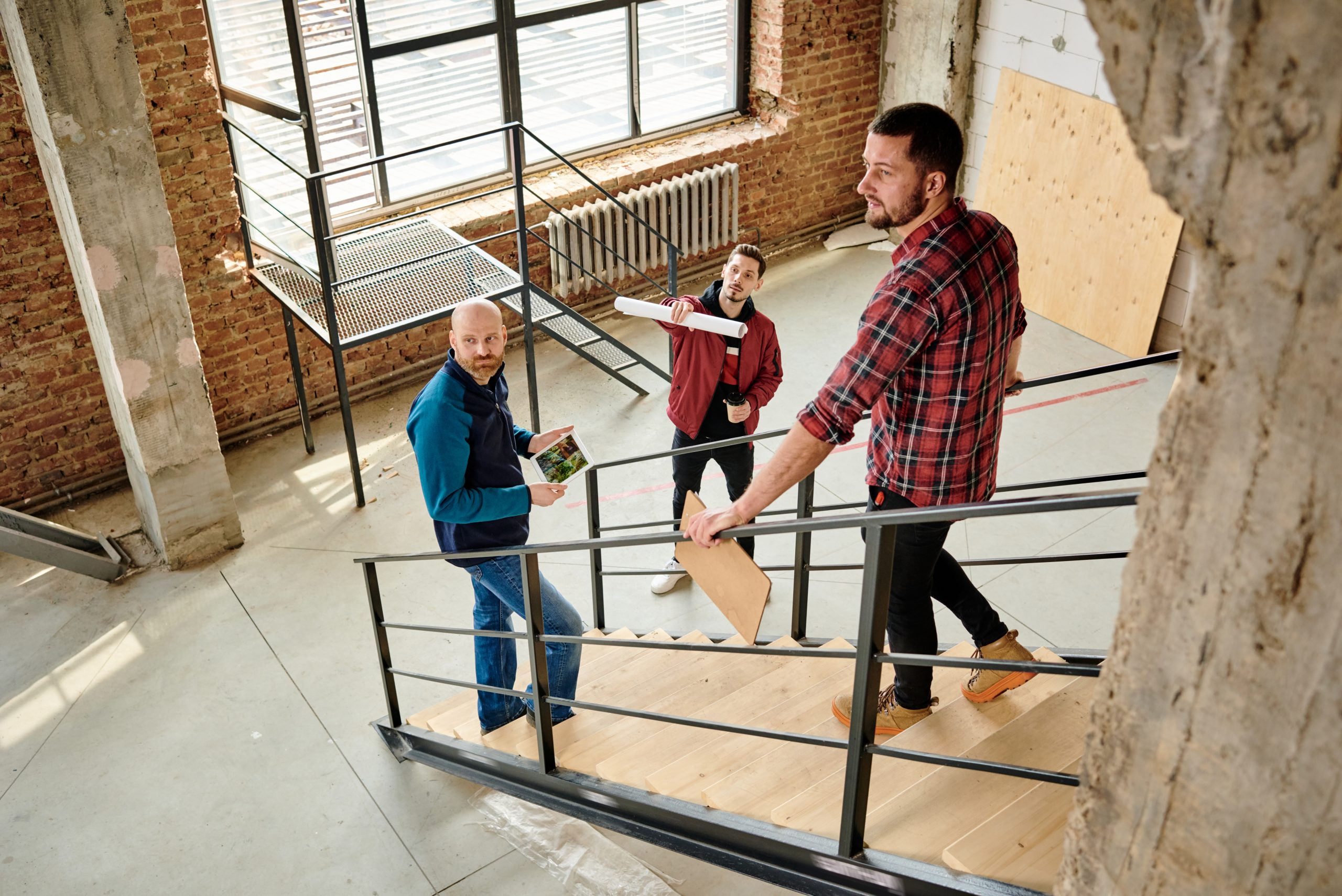 Young architect with rolled blueprint in hand pointing at wall while standing by staircase and discussing details of quality with colleagues