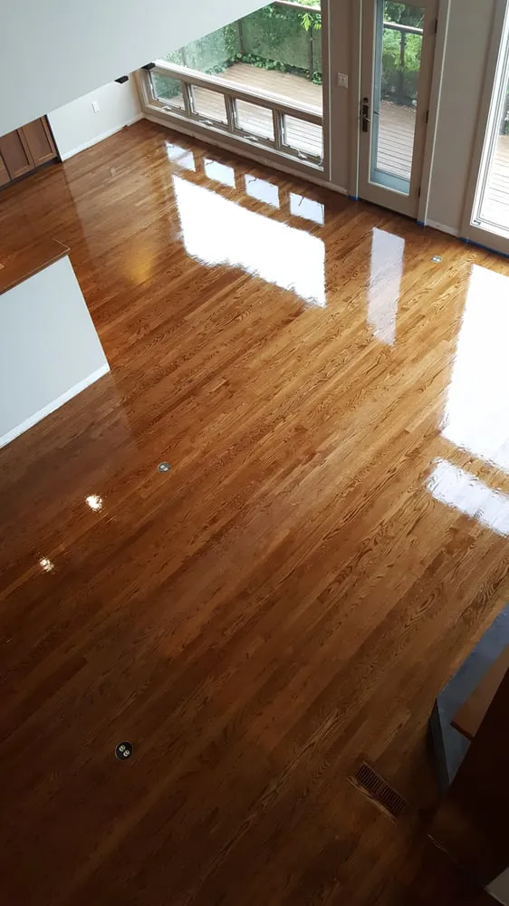 High-angle view of a high-gloss refinished hardwood floor in an open-concept living space with large windows and a sunlit reflection.