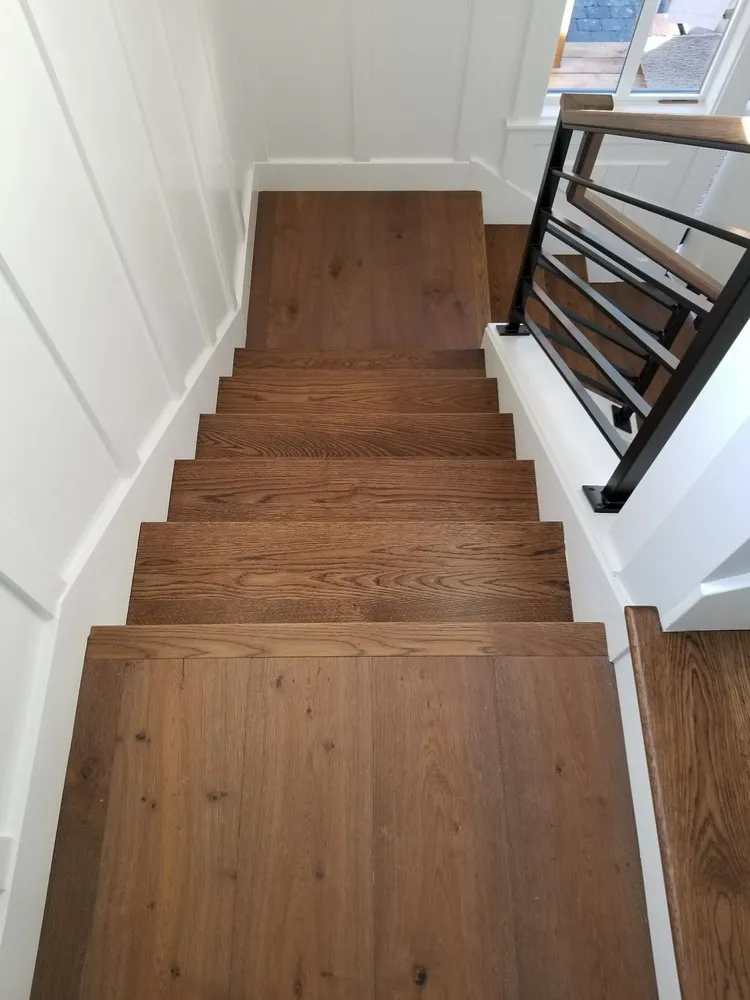 Top-down view of modern hardwood stairs with wide planks, white shiplap walls, and a black and wood railing